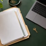 Dossier de documents près d’un ordinateur portable et d’une petite clé sur un bureau vert.